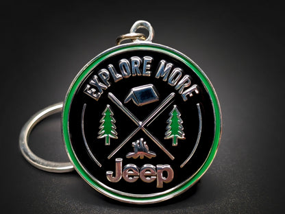 Jeep "Explore More" badge keychain featuring 3D camping icons, pine trees, and a green enamel border on a black background.