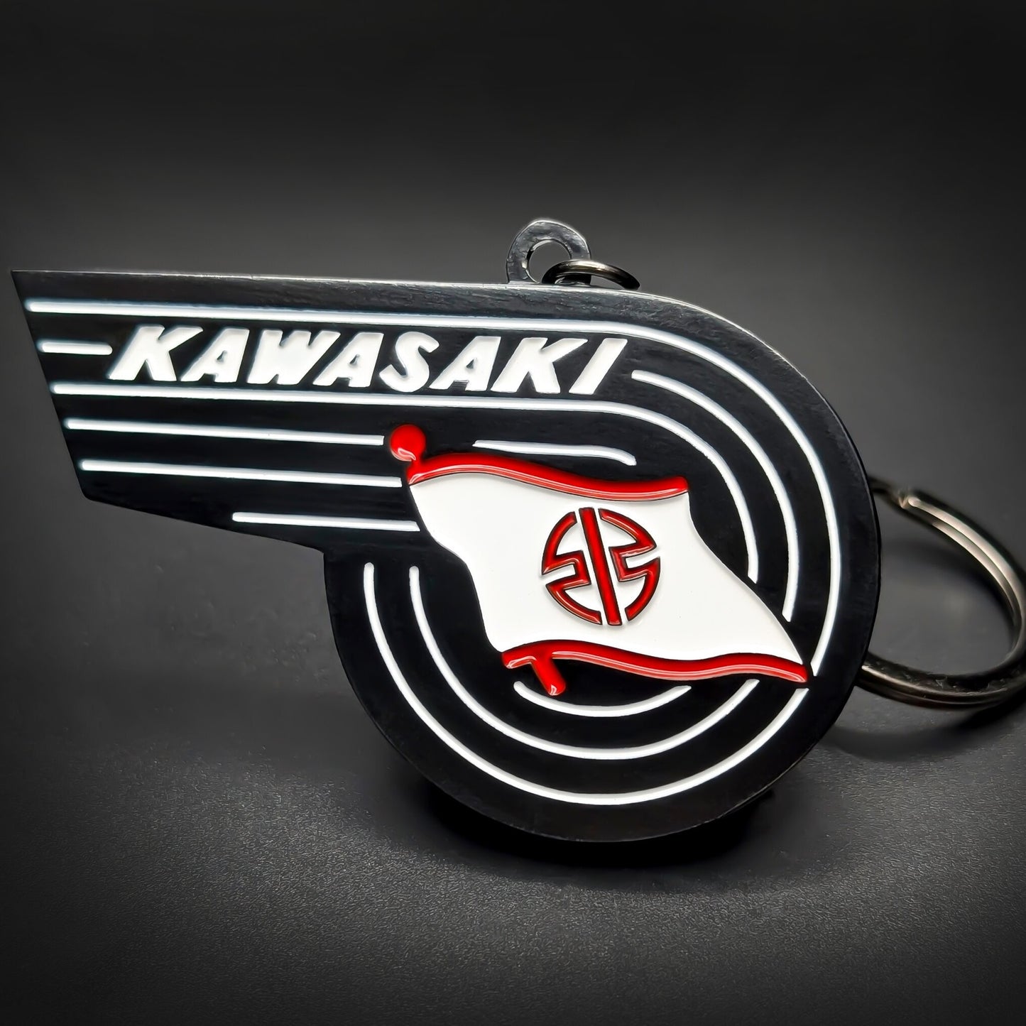 Double-sided metal Kawasaki badge keychain shaped like a swept wing, with white ‘KAWASAKI’ lettering and a red-and-white flag emblem on black enamel, chrome split ring attached, on a dark surface.