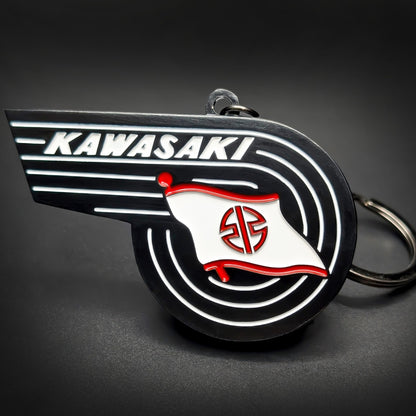 Double-sided metal Kawasaki badge keychain shaped like a swept wing, with white ‘KAWASAKI’ lettering and a red-and-white flag emblem on black enamel, chrome split ring attached, on a dark surface.