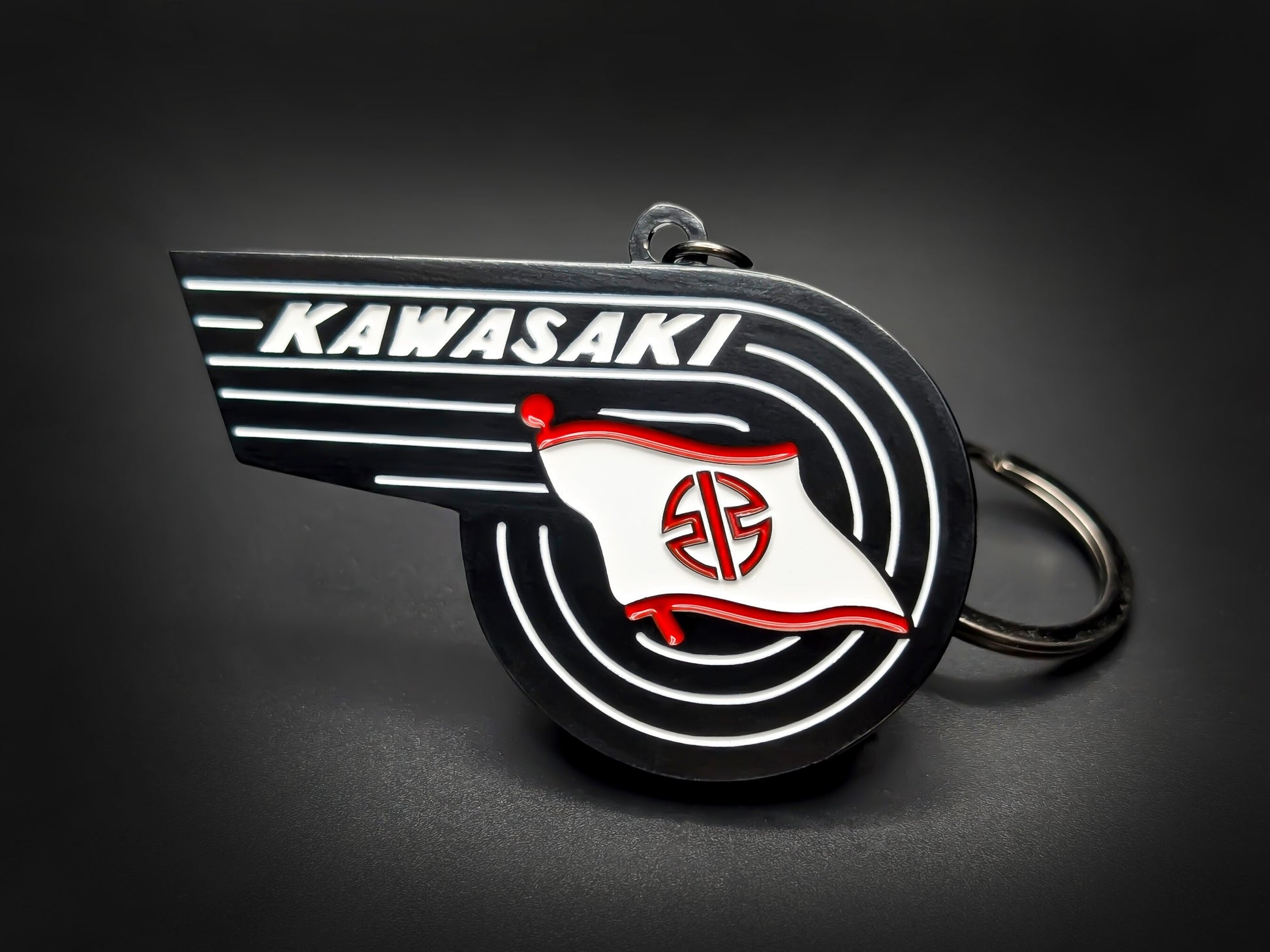 Vintage Kawasaki badge keychain featuring the winged wordmark and white River Mark flag with red accents on black enamel.