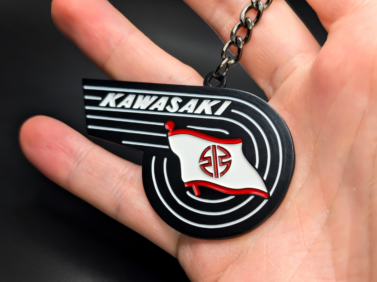 Hand holding the Kawasaki heritage badge keychain to display the 3D raised details and premium matte black finish.