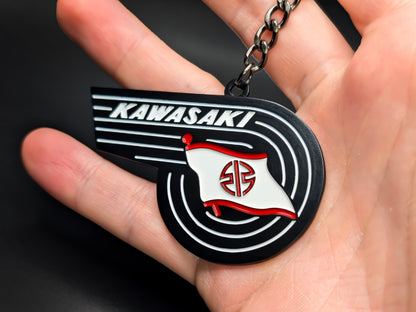 Hand holding the Kawasaki heritage badge keychain to display the 3D raised details and premium matte black finish.