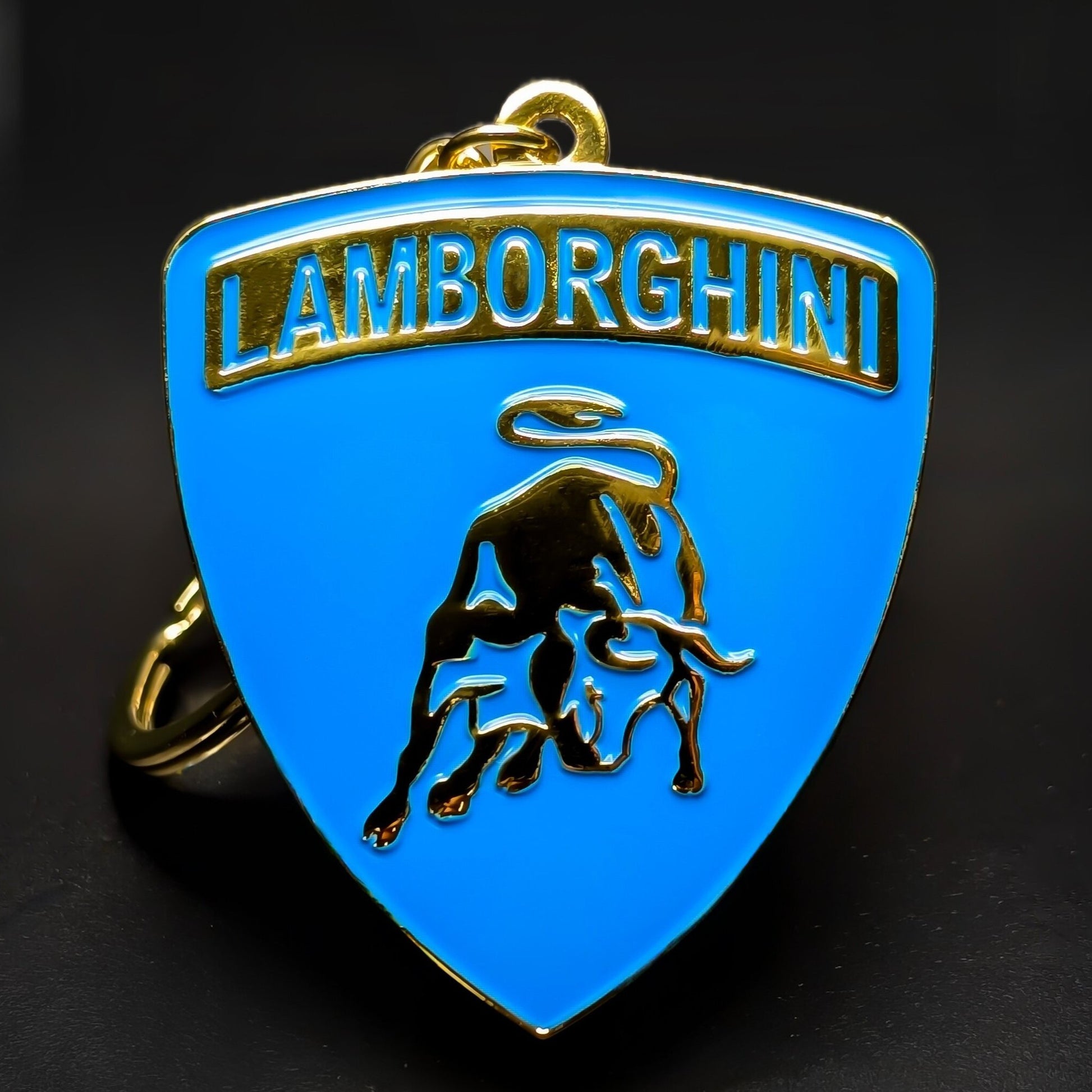 Double-sided metal Lamborghini badge keychain in blue enamel with gold trim, blue LAMBORGHINI lettering, and a gold bull emblem, hanging from a gold split ring on a dark surface.