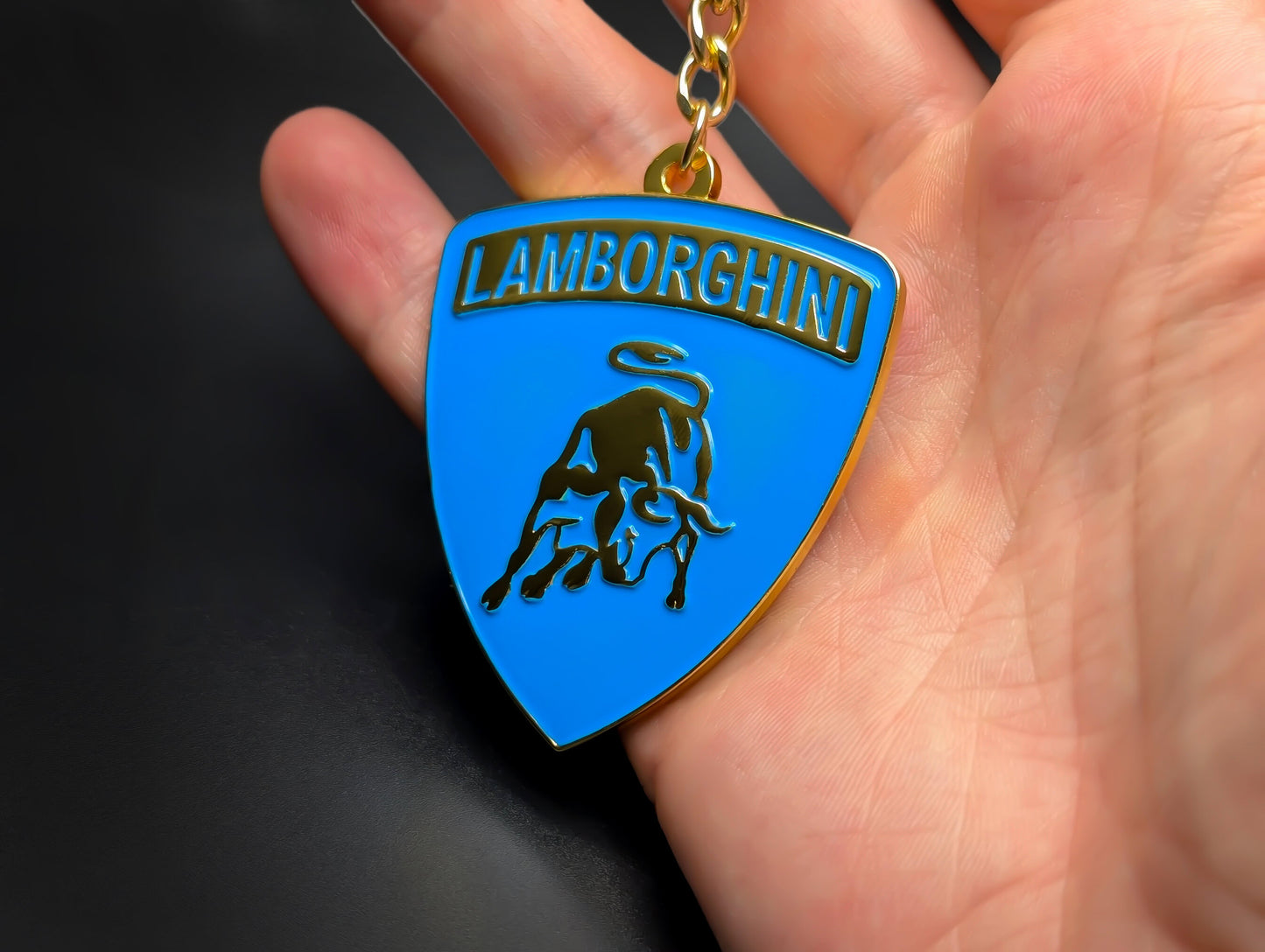 Hand holding a blue and gold Lamborghini enamel badge keychain to demonstrate the metallic weight and premium finish.
