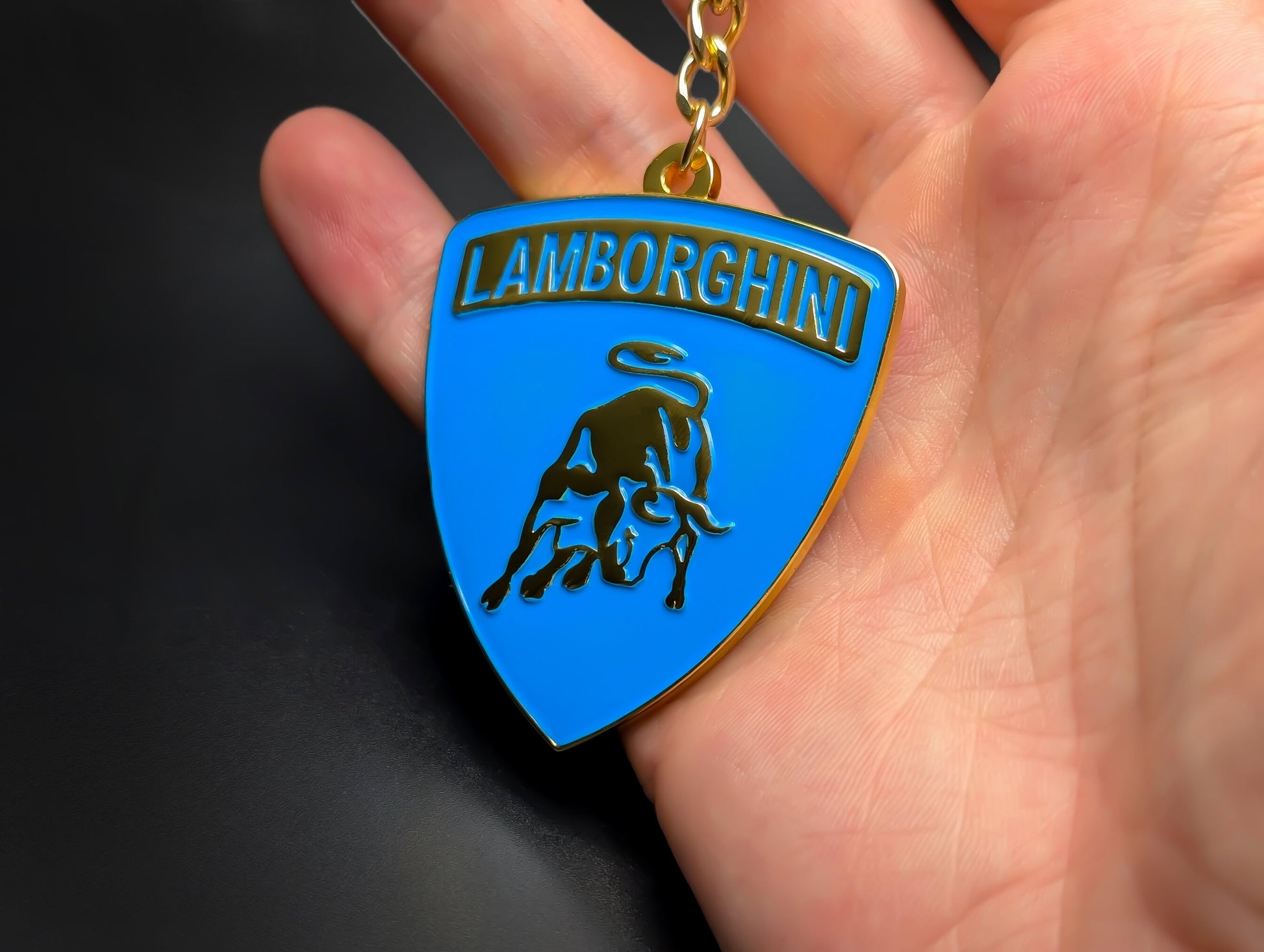 Hand holding a blue and gold Lamborghini enamel badge keychain to demonstrate the metallic weight and premium finish.