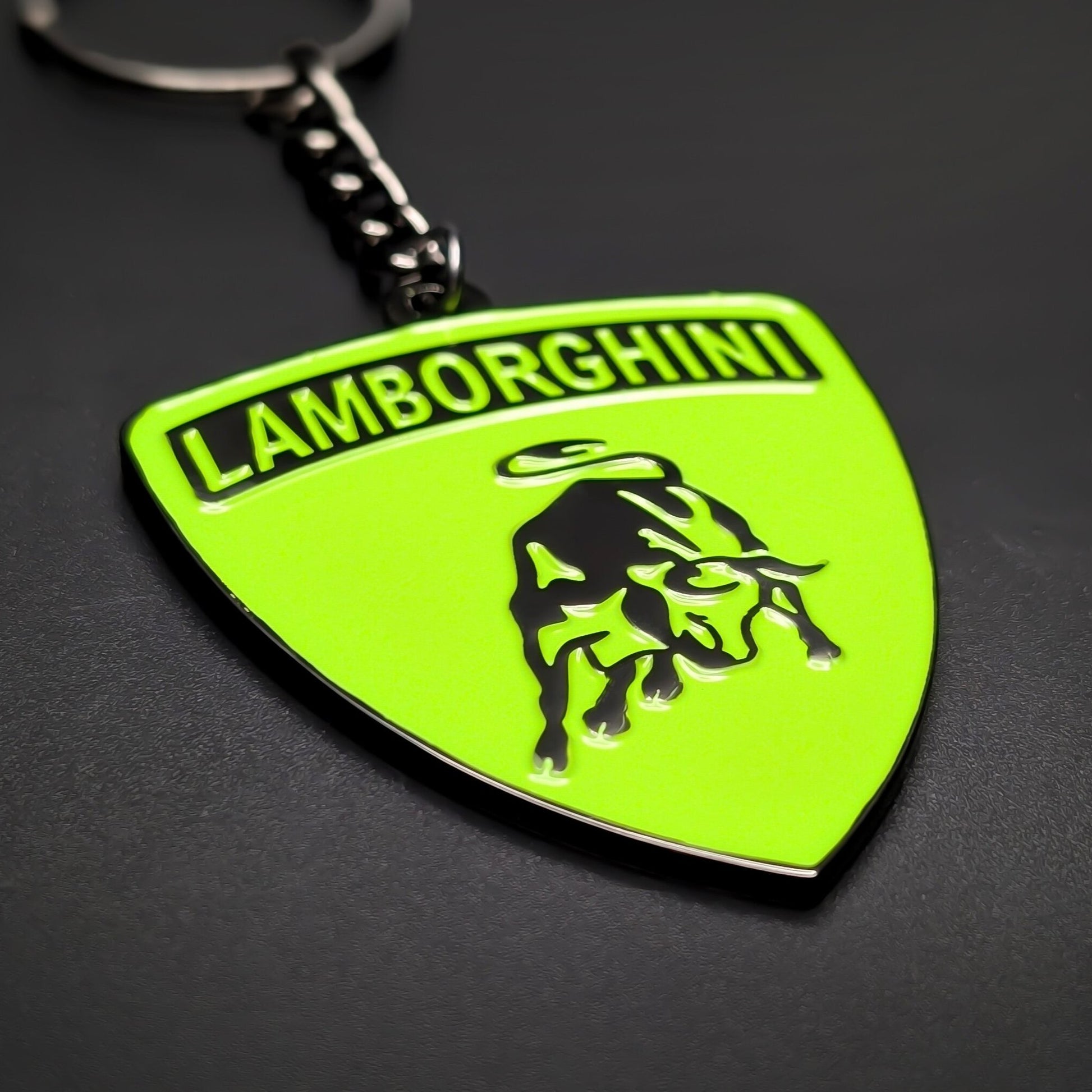 Double-sided metal Lamborghini badge keychain in lime green enamel with black LAMBORGHINI lettering and a black bull emblem, attached to a split ring by a chain, on a dark textured surface.