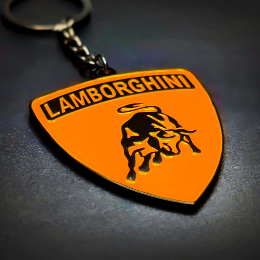 Double-sided metal Lamborghini badge keychain in orange enamel with black LAMBORGHINI lettering and a black bull emblem, attached to a split ring by a chain, on a dark textured surface.