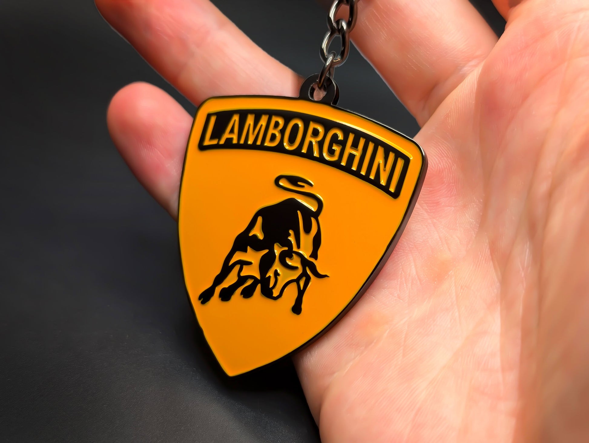 Hand holding an orange and black Lamborghini enamel badge keychain to demonstrate the premium weight and metallic finish.