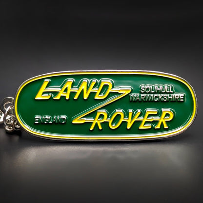 Double-sided oblong metal Land Rover badge keychain, green enamel face with a yellow LAND ROVER wordmark, ENGLAND and SOLIHULL WARWICKSHIRE lettering in white, chrome trim, chain and split ring, on a dark surface.
