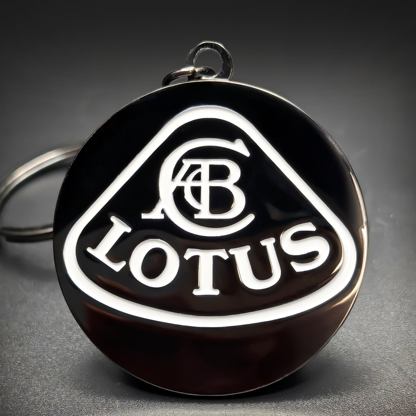 Double-sided metal Lotus badge keychain with a white triangular shield outline, an interwoven monogram, and ‘LOTUS’ lettering on black enamel, chrome split ring attached, on a dark surface.