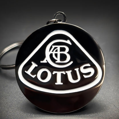 Double-sided metal Lotus badge keychain with a white triangular shield outline, an interwoven monogram, and ‘LOTUS’ lettering on black enamel, chrome split ring attached, on a dark surface.