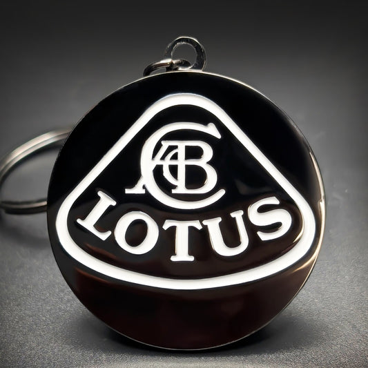 Double-sided metal Lotus badge keychain with a white triangular shield outline, an interwoven monogram, and ‘LOTUS’ lettering on black enamel, chrome split ring attached, on a dark surface.