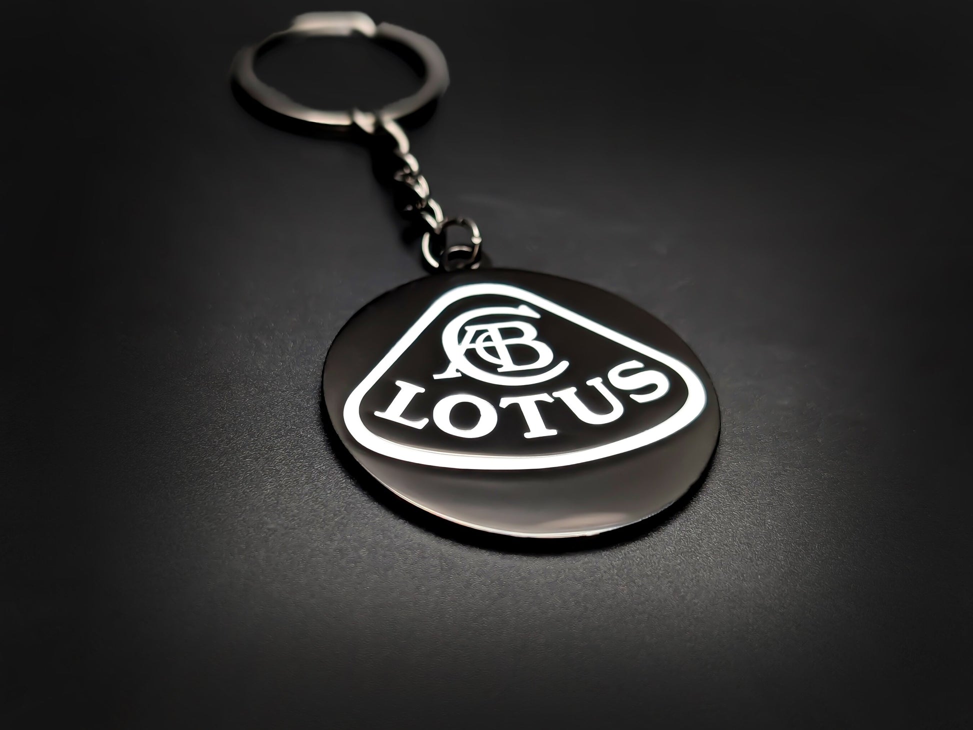 High-gloss black Lotus badge keychain on a dark background, showing the polished silver-tone chain and circular metal base.