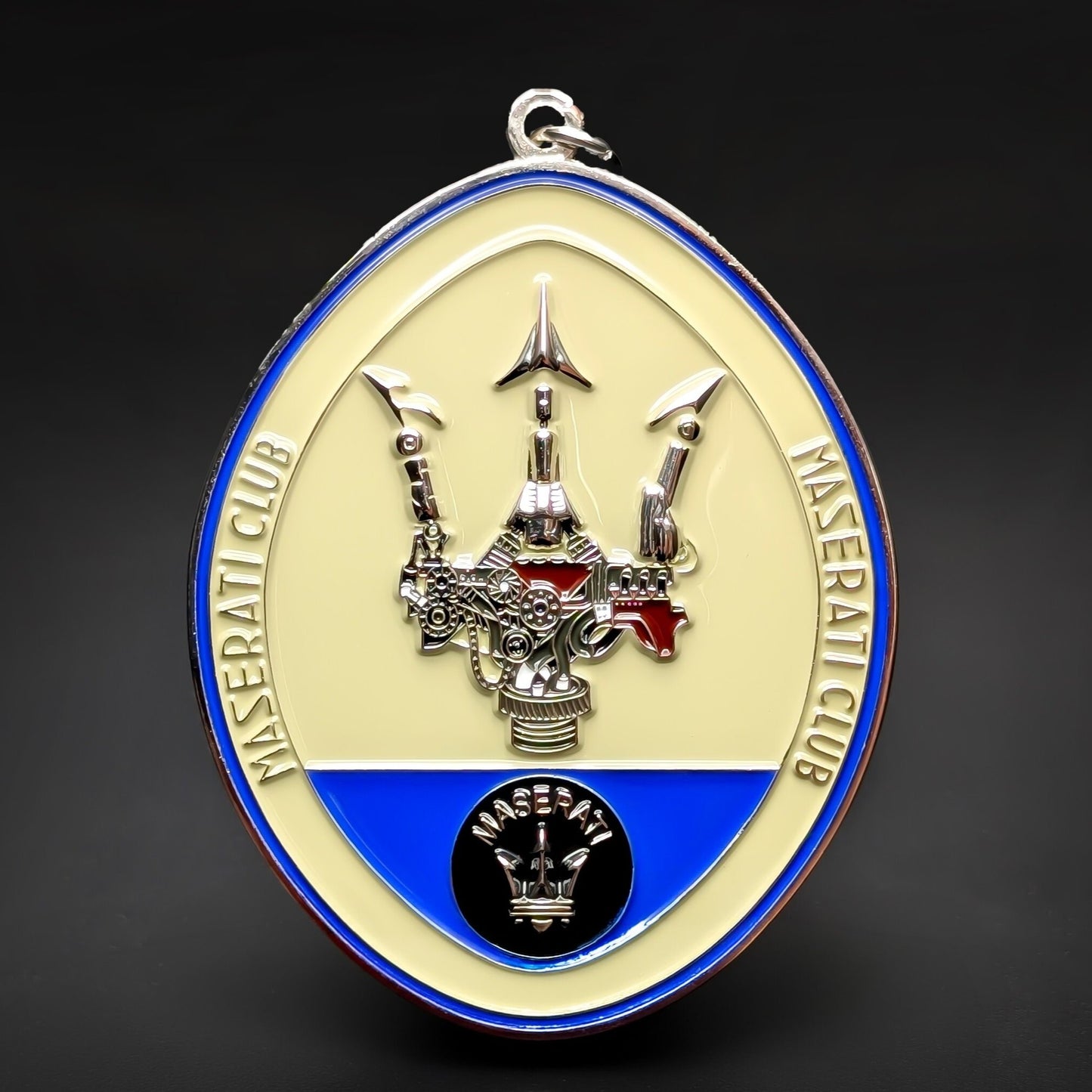 Double-sided oval metal Maserati Club badge keychain, cream face and blue ring reading MASERATI CLUB, a chrome trident over a silver-and-red engine, black MASERATI emblem below, chrome border and hanging loop.