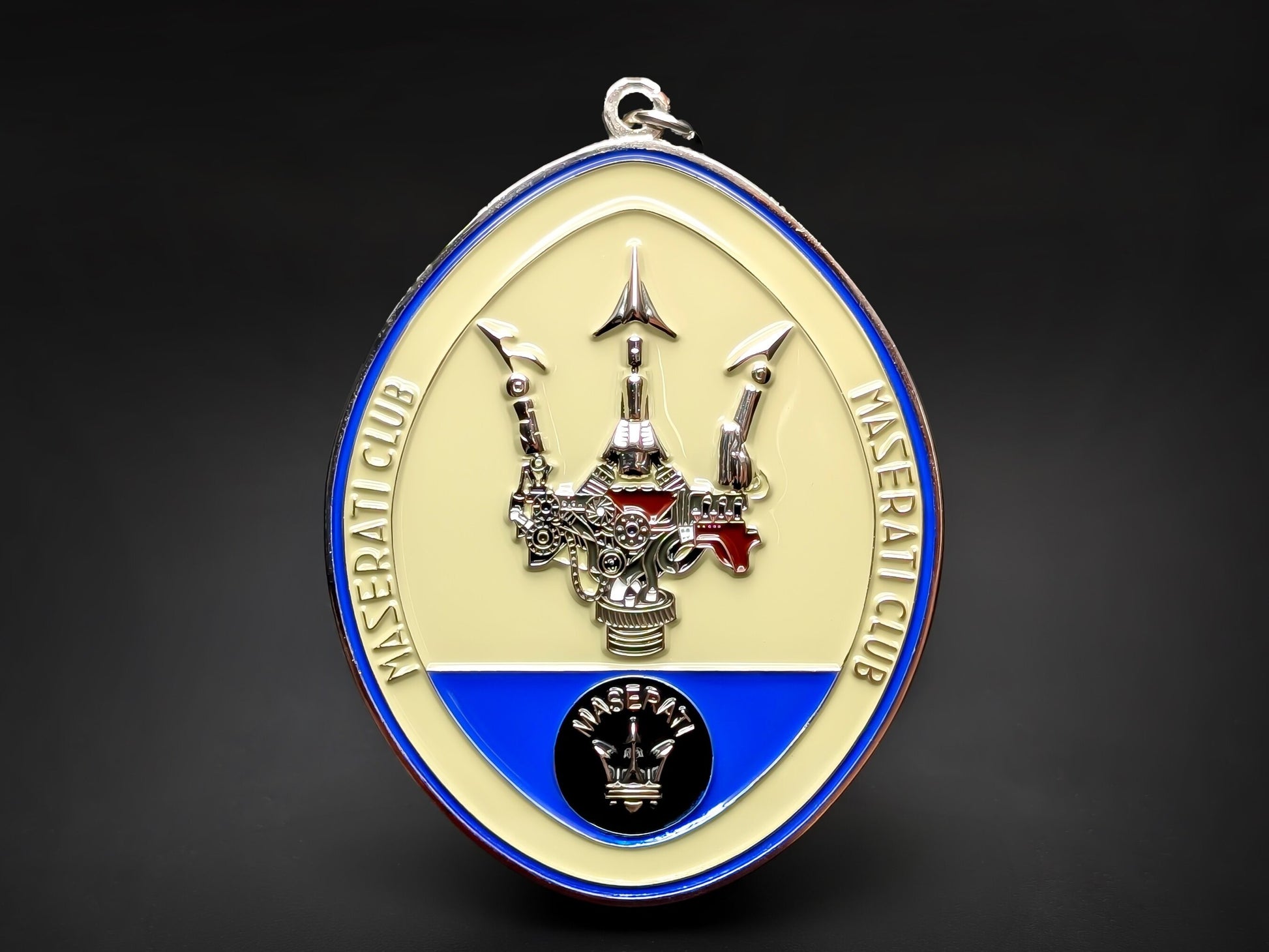 Maserati Club badge keychain featuring a mechanical-style silver trident on a cream and blue oval enamel background.