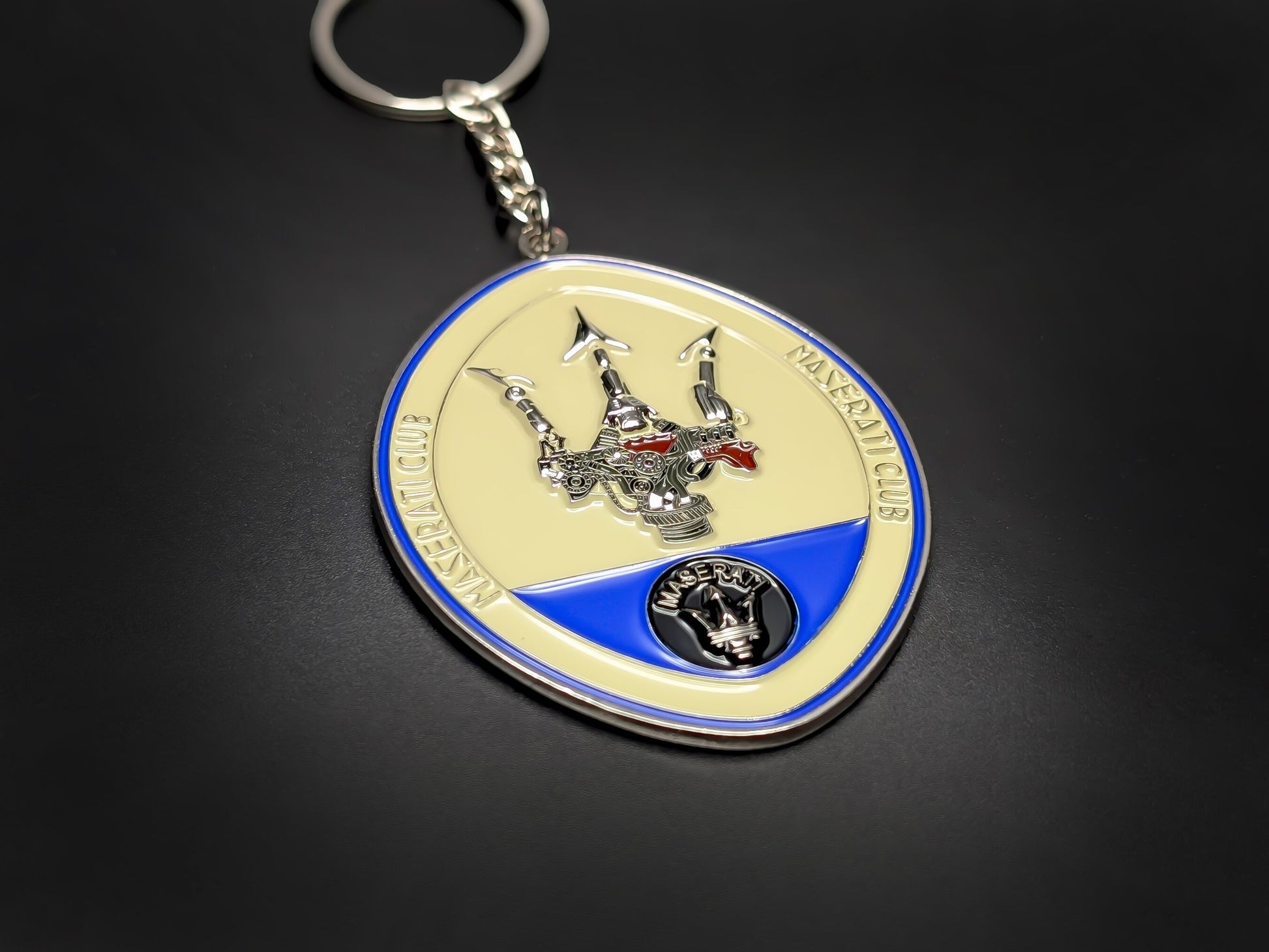 High-gloss Maserati car club badge keychain on a dark background, showing the polished silver metal and blue enamel trim.
