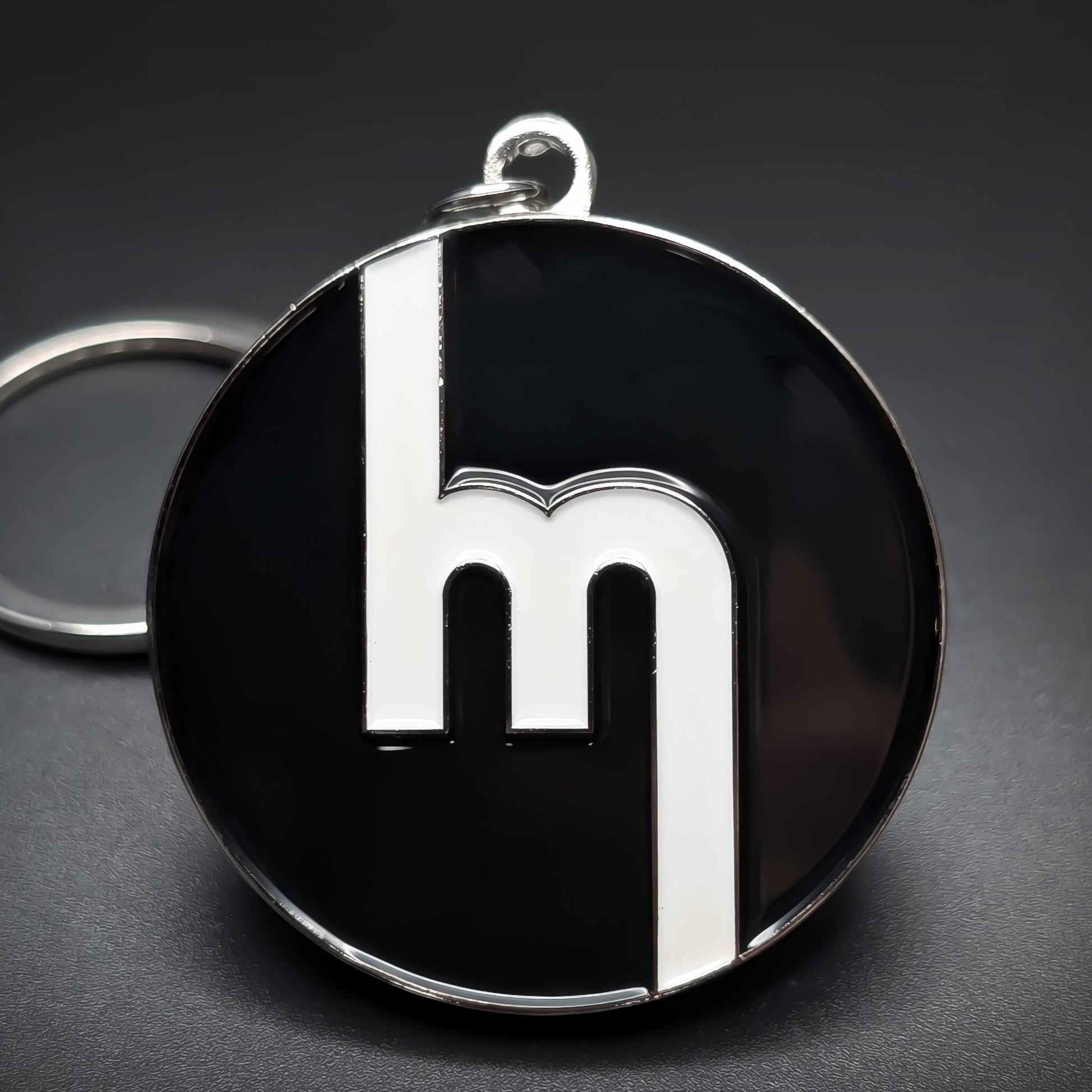 Double-sided metal Mazda badge keychain, a round chrome-rimmed medallion in glossy black enamel with a white stylized lowercase m mark centered, split ring attached, on a dark surface.