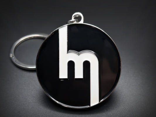 Vintage Mazda "M" badge keychain featuring the classic white block logo on a circular black enamel base.