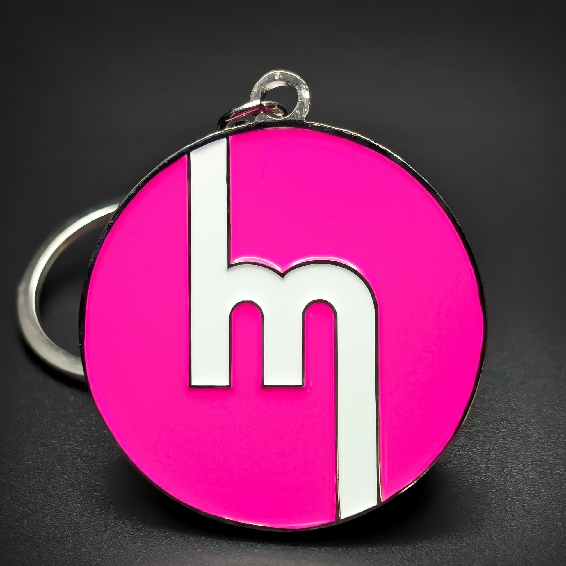 Double-sided metal Mazda badge keychain, a round chrome-rimmed medallion in glossy pink enamel with a white stylized lowercase m mark centered, split ring attached, on a dark surface.