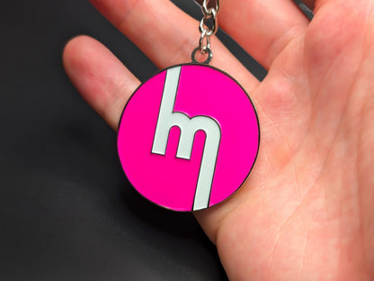 Hand holding a circular neon pink Mazda heritage enamel badge keychain to demonstrate the high-contrast design and premium finish.