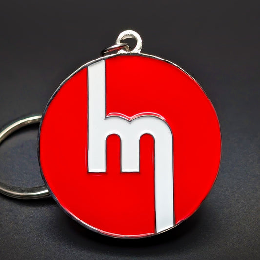 Double-sided metal Mazda badge keychain, a round chrome-rimmed medallion in glossy red enamel with a white stylized lowercase m mark centered, split ring attached, on a dark surface.