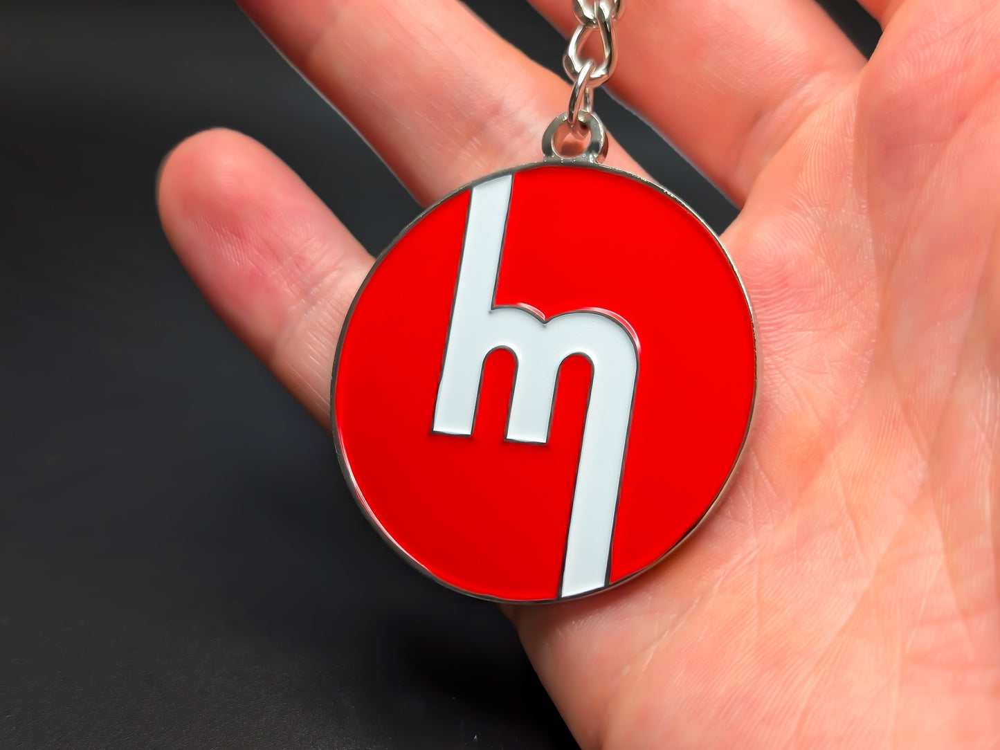 Circular red Mazda heritage enamel badge keychain held in hand to demonstrate the premium metallic weight and vibrant color finish.