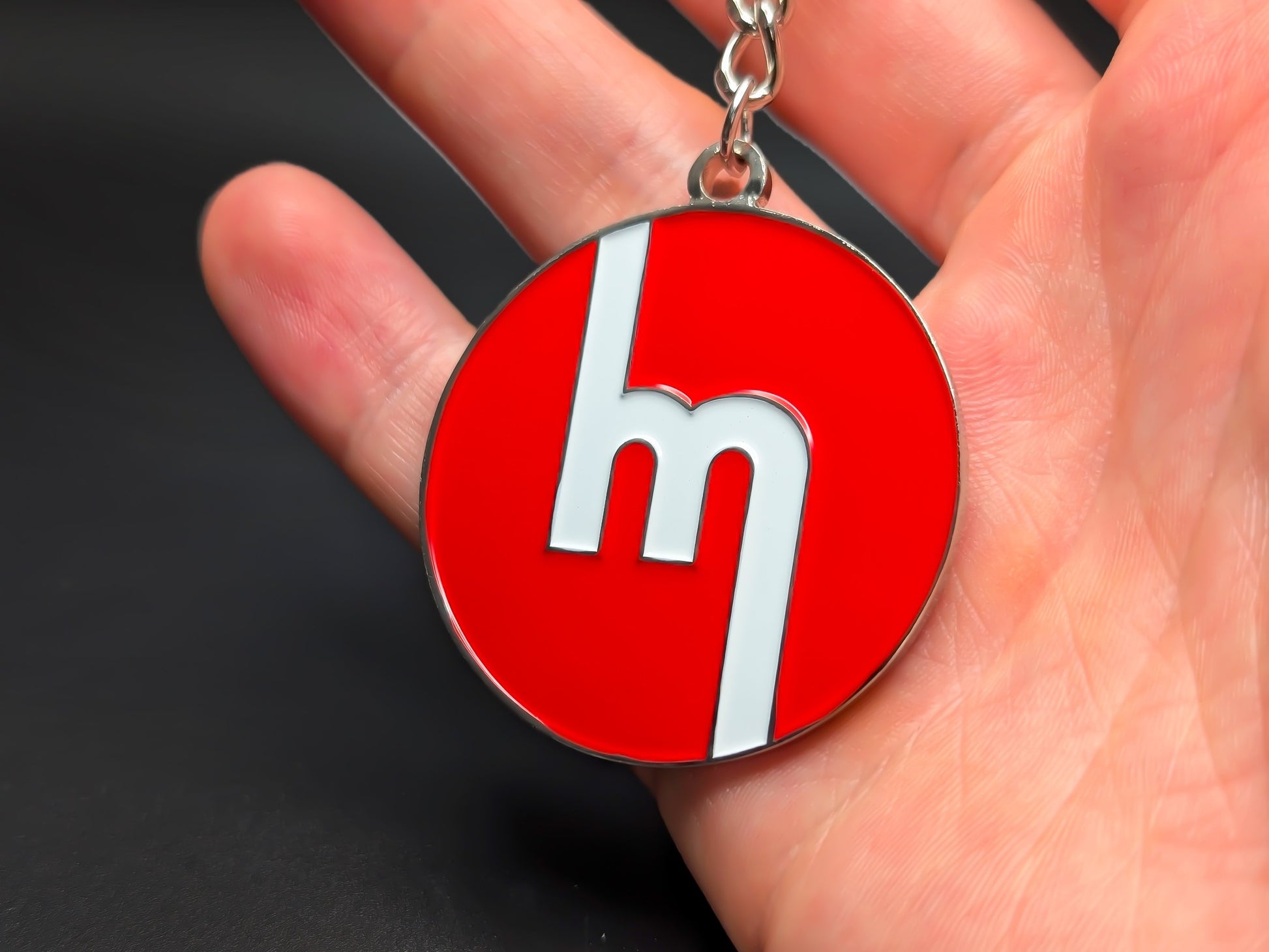 Circular red Mazda heritage enamel badge keychain held in hand to demonstrate the premium metallic weight and vibrant color finish.