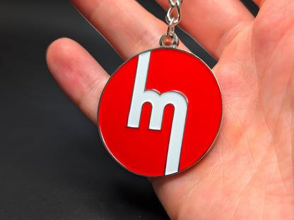Circular red Mazda heritage enamel badge keychain held in hand to demonstrate the premium metallic weight and vibrant color finish.