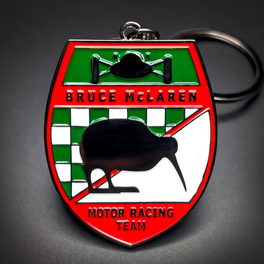 Double-sided metal Bruce McLaren badge keychain, red, green, and white enamel shield with a race car graphic, “Bruce McLaren” and “Motor Racing Team” text, a black kiwi over checkers, split ring.