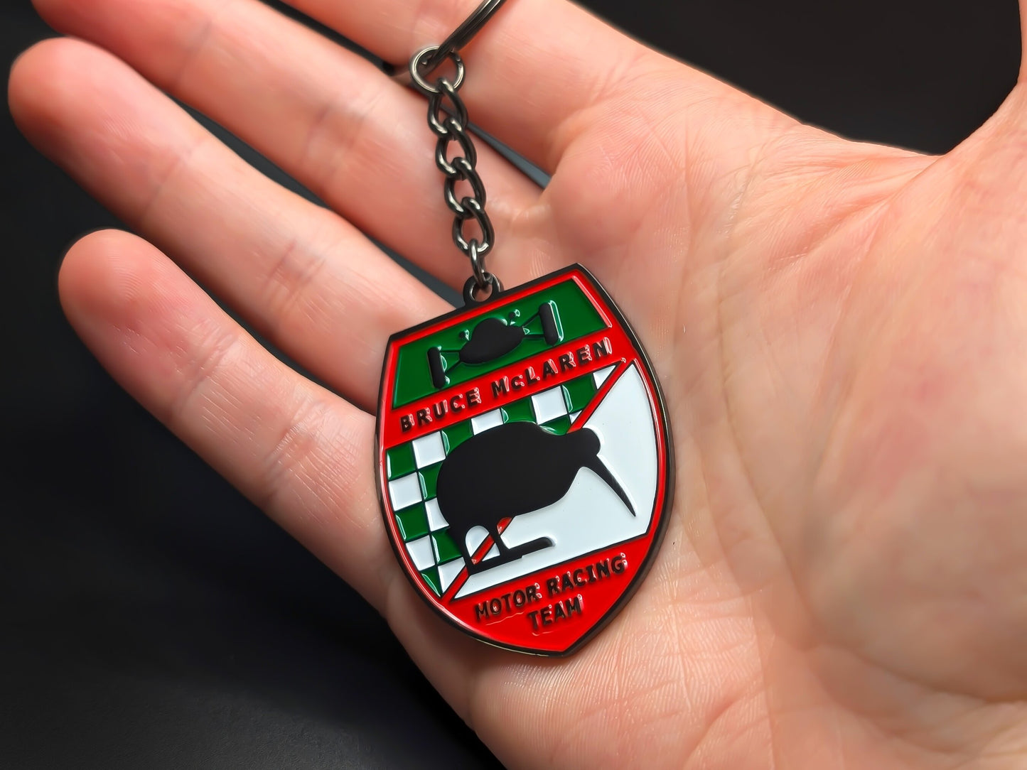 Hand holding a shield-shaped Bruce McLaren enamel badge keychain to demonstrate the premium metallic weight and detailed 3D color finish.