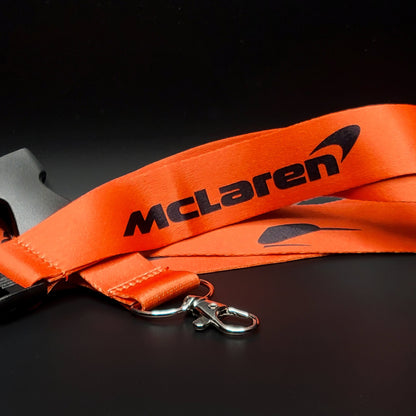 Orange McLaren lanyard with a black McLaren logo and the black Speedy Kiwi bird emblem repeated along the strap, coiled with a black breakaway clip and silver clasp on black.