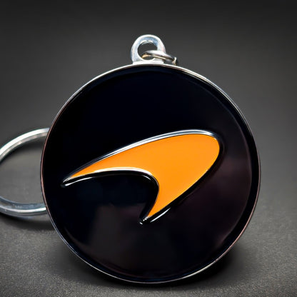 Double-sided metal McLaren badge keychain, a black circular badge with a papaya-orange speedmark swoosh at center and a chrome rim, on a jump ring against a dark background.