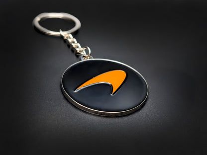 High-quality McLaren racing badge keychain on a dark background, showing the polished silver-tone metal chain and rim.