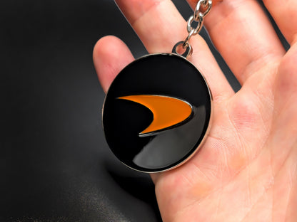 Hand holding a circular McLaren enamel badge keychain to demonstrate the vibrant orange color and premium metallic finish.