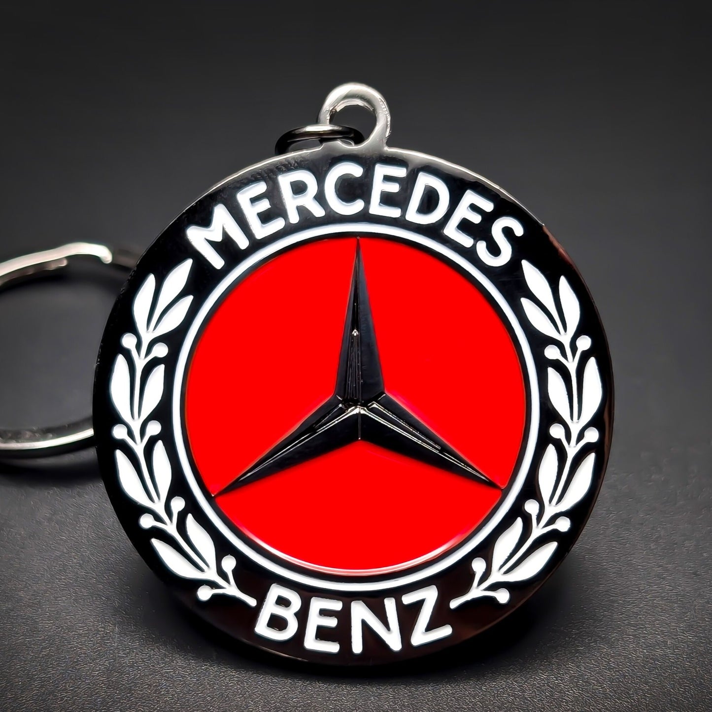 Double-sided metal Mercedes-Benz badge keychain, black roundel with white ‘MERCEDES’ and ‘BENZ’ lettering, laurel branches, red center, and black three-pointed star, on a split ring on a dark background.