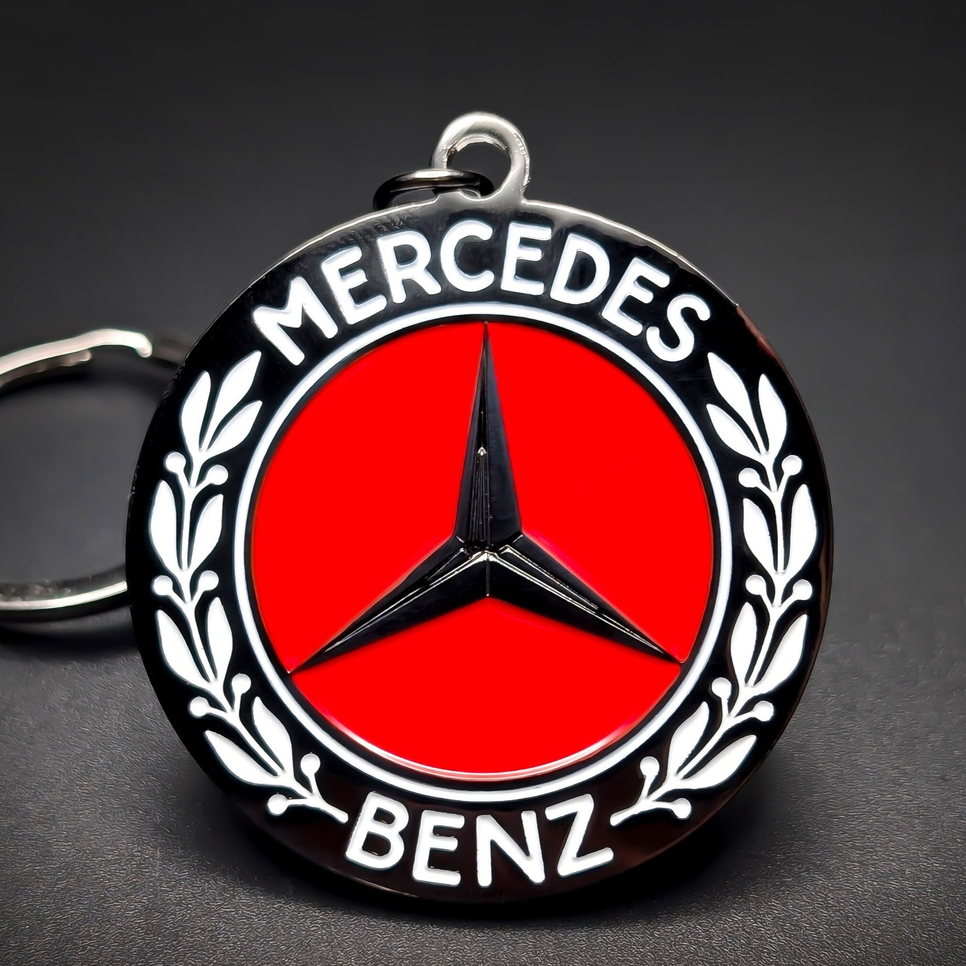 Double-sided metal Mercedes-Benz badge keychain, black roundel with white ‘MERCEDES’ and ‘BENZ’ lettering, laurel branches, red center, and black three-pointed star, on a split ring on a dark background.