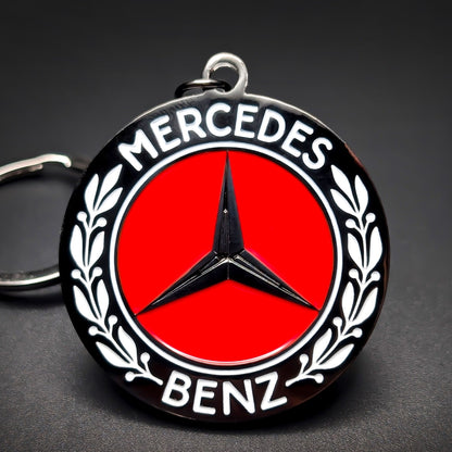 Double-sided metal Mercedes-Benz badge keychain, black roundel with white ‘MERCEDES’ and ‘BENZ’ lettering, laurel branches, red center, and black three-pointed star, on a split ring on a dark background.