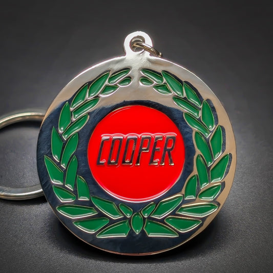  Double-sided metal Mini Cooper badge keychain, a round chrome medallion with a green laurel wreath framing a red center disc reading COOPER in black lettering, split ring, on a dark surface.