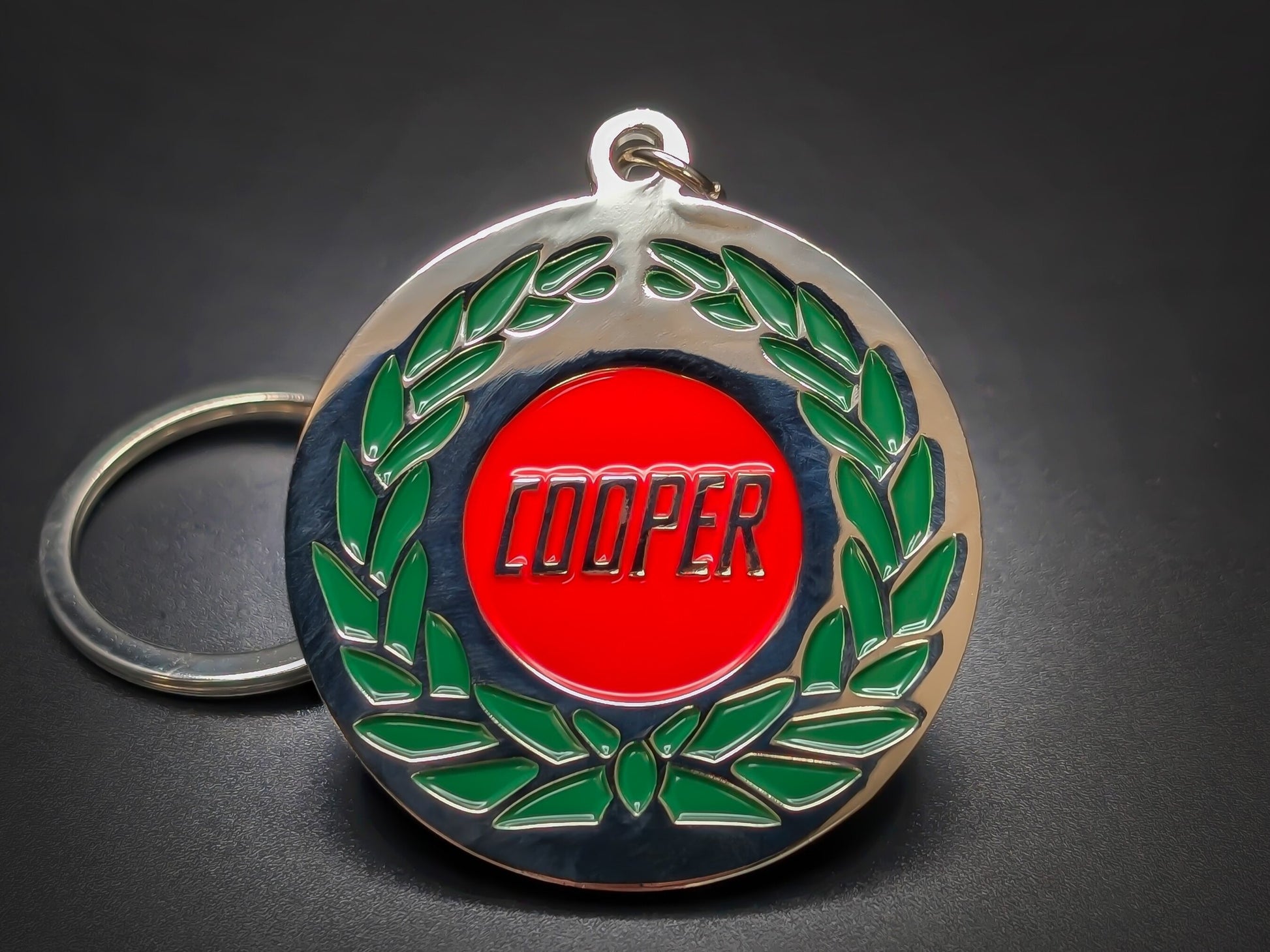 Mini Cooper badge keychain featuring a red "COOPER" center surrounded by a green enamel laurel wreath on a polished silver base.
