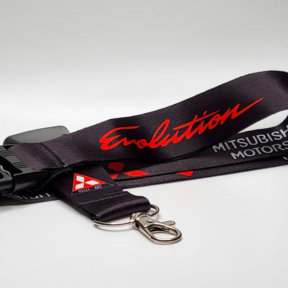 Black Mitsubishi lanyard with red script Evolution text, red three-diamond logos, and a red-and-white Ralli Art triangle badge, coiled with a black breakaway clip and silver clasp on white.