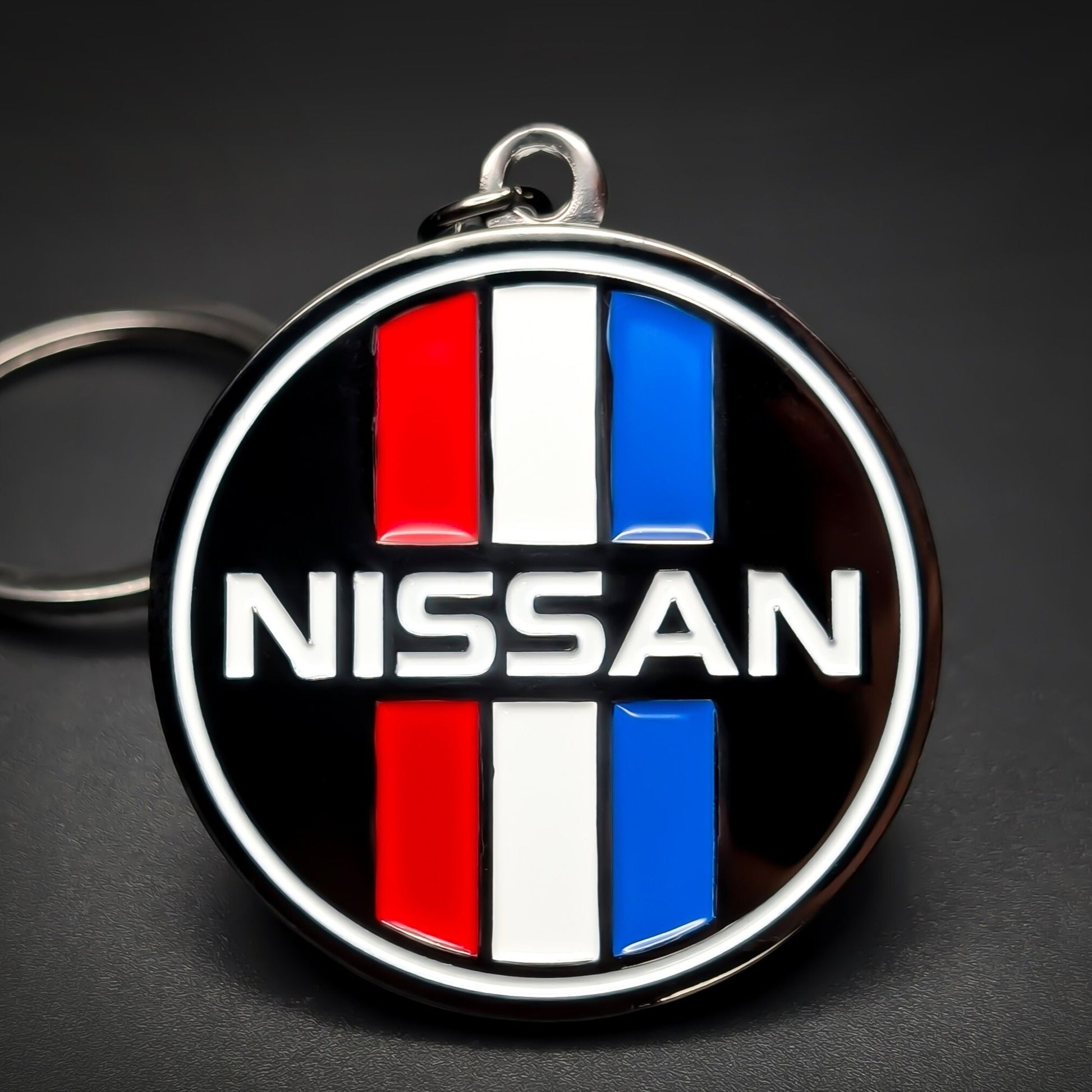 Double-sided metal Nissan badge keychain, a black badge with red, white, and blue stripes above and below ‘NISSAN’ in white block letters, chrome rim, on a split ring against a dark background.
