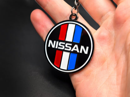 Circular Nissan heritage badge keychain held in hand, displaying the high-contrast racing colors and premium 3D raised enamel finish.
