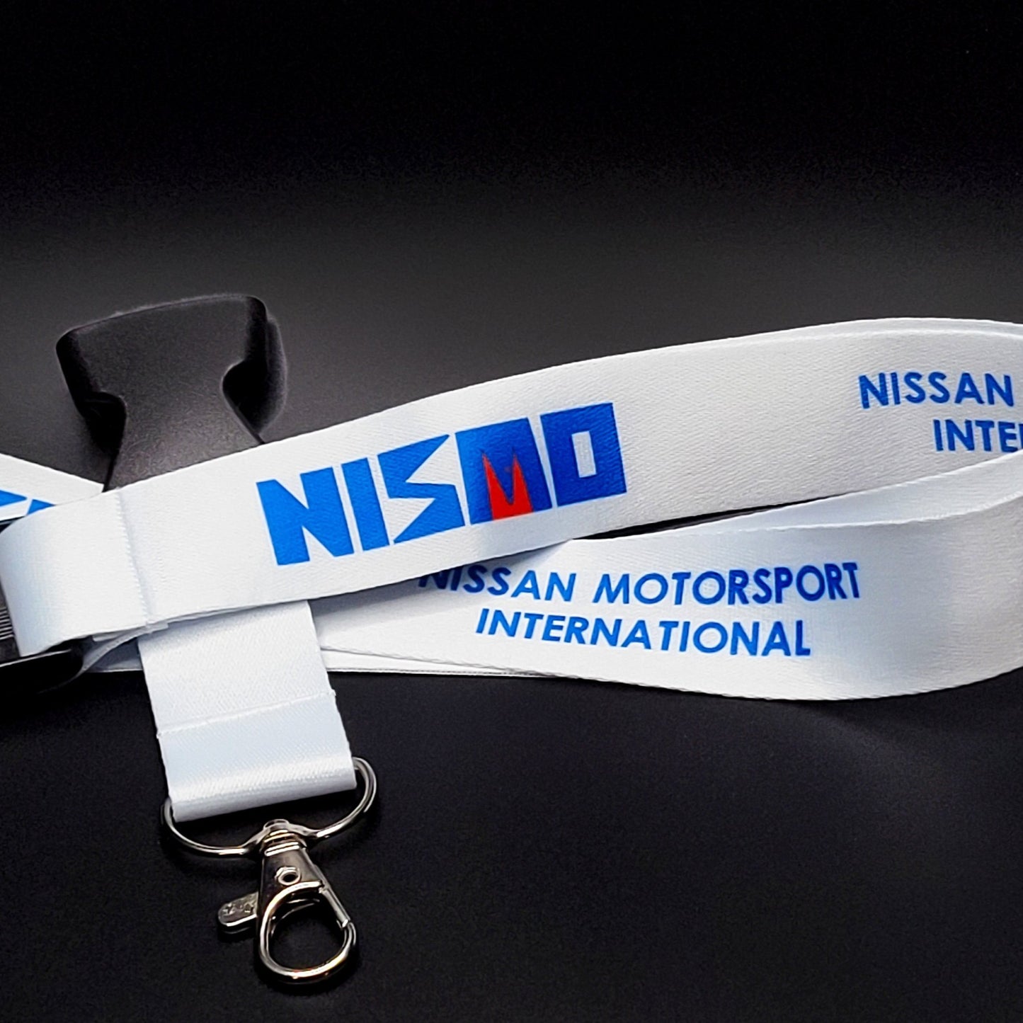 White Nissan Nismo lanyard with a blue-and-red NISMO logo and blue Nissan Motorsport International text, coiled with a black breakaway clip and silver clasp on black.