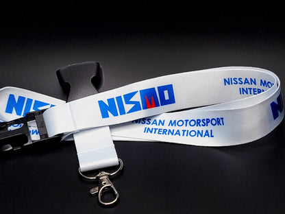 White Nissan Nismo lanyard featuring blue Nismo block lettering with a red accent, silver metal clasp, and black quick-release buckle.