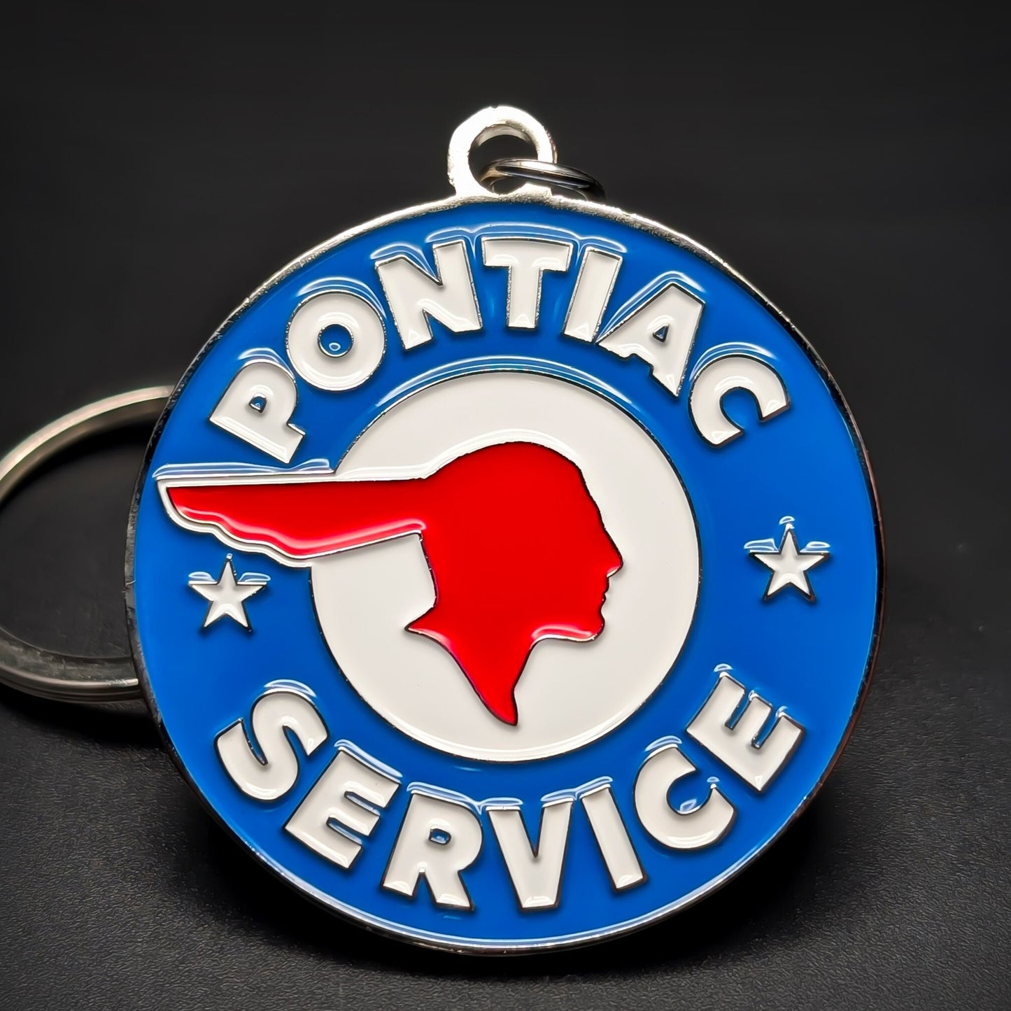 Double-sided metal Pontiac Service badge keychain, a blue medallion with a red chief-head emblem in a white circle, PONTIAC and SERVICE in white lettering, a star each side, dark surface.