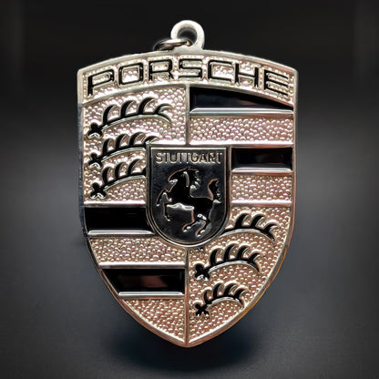 Double-sided metal Porsche crest badge keychain in silver with black enamel bands, a central STUTTGART shield with a black horse, textured quarters, and a small top loop, on a dark surface.