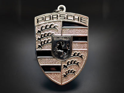 Porsche black badge keychain with silver 3D crest, black enamel stripes, and Stuttgart horse on a textured metallic shield.