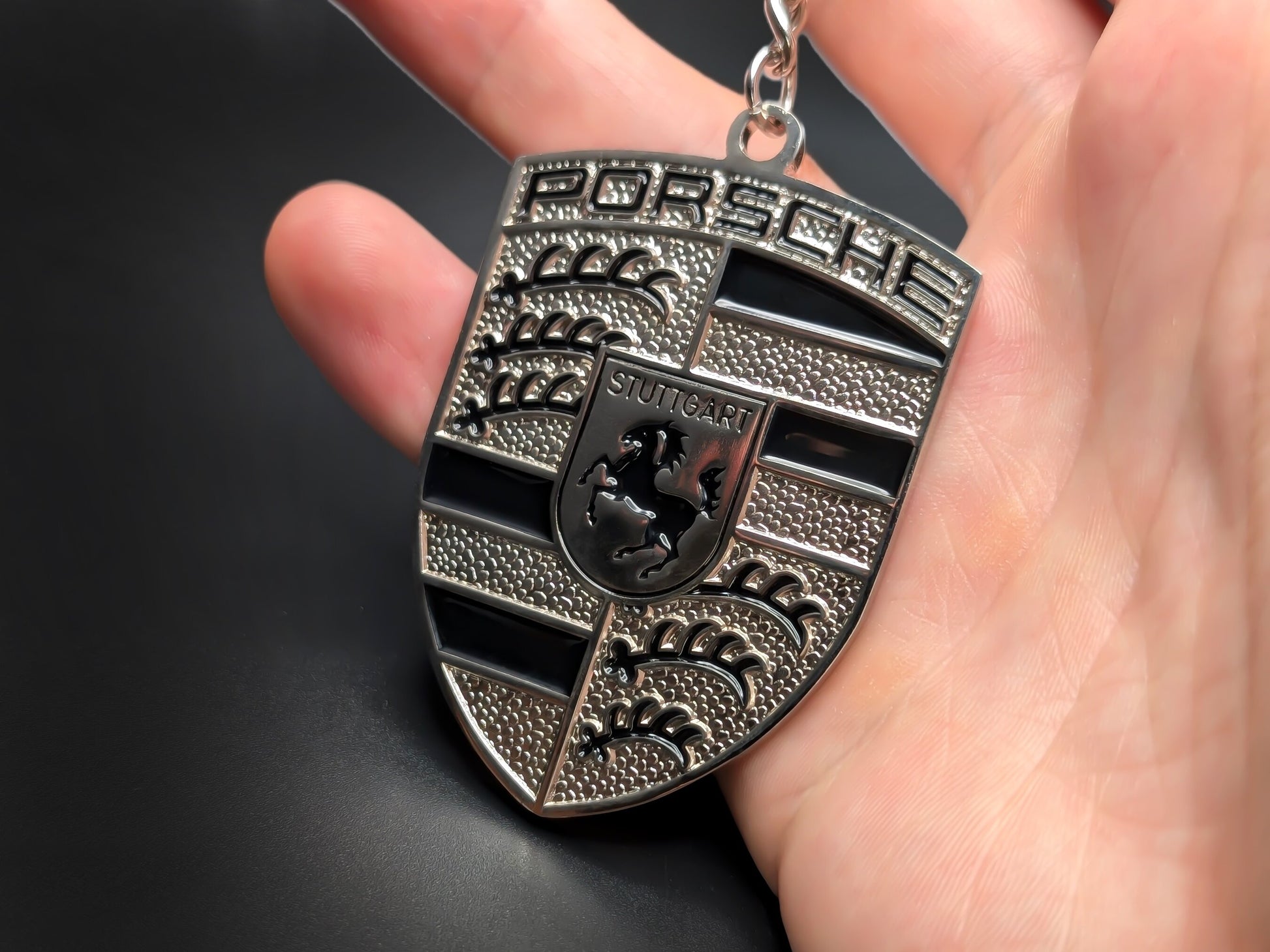 Hand holding a monochrome black Porsche enamel badge keychain to demonstrate the premium metallic weight and 3D textures.