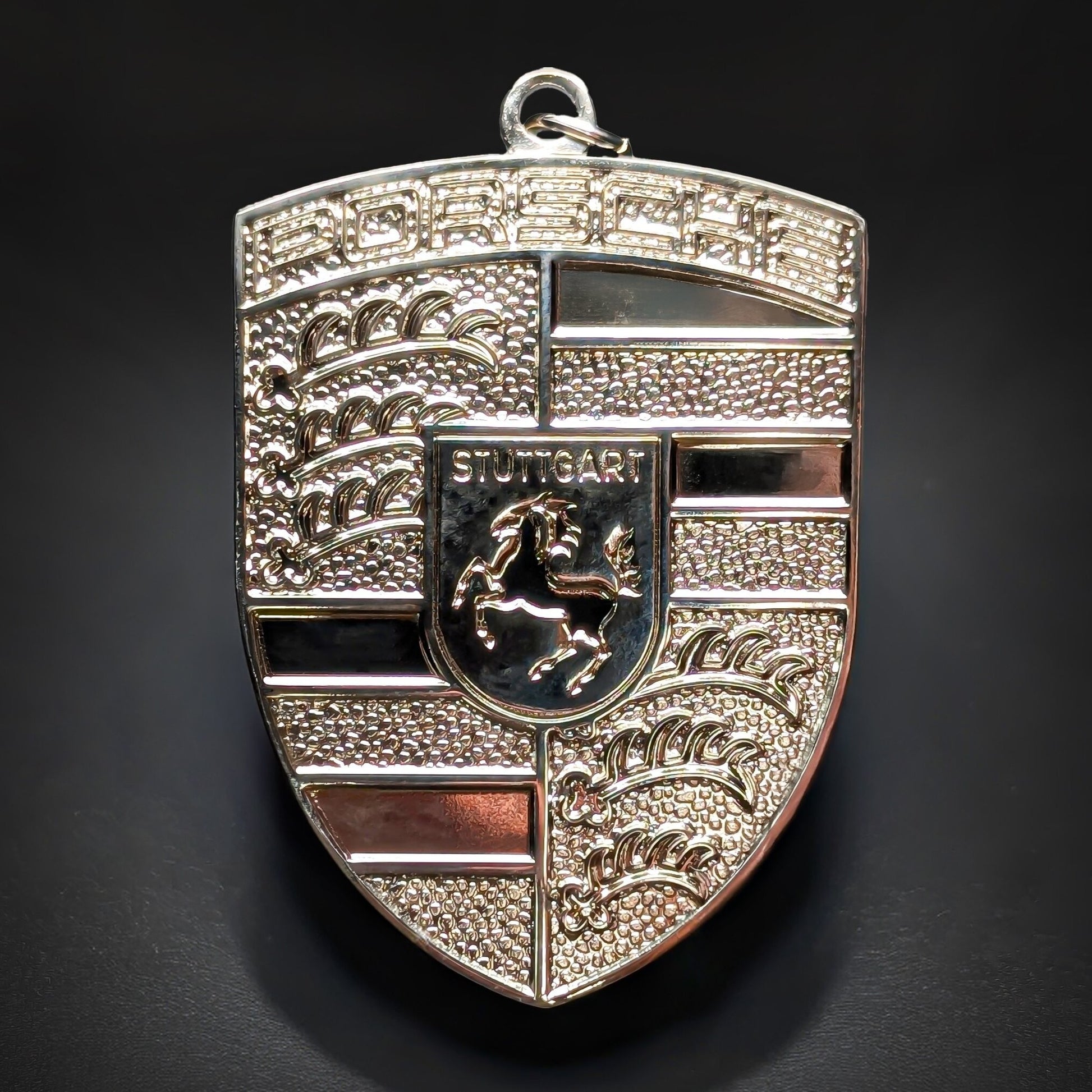 Double-sided metal Porsche crest badge keychain in bright polished silver with black enamel bands, a central STUTTGART shield with a black horse, and a reflective PORSCHE wordmark, on a dark surface.