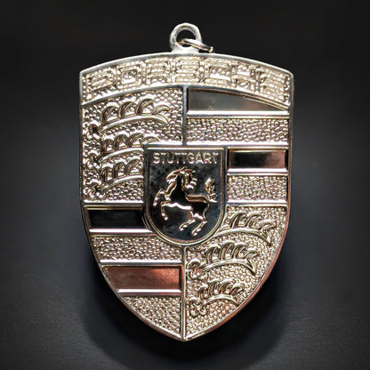 Double-sided metal Porsche crest badge keychain in bright polished silver with black enamel bands, a central STUTTGART shield with a black horse, and a reflective PORSCHE wordmark, on a dark surface.